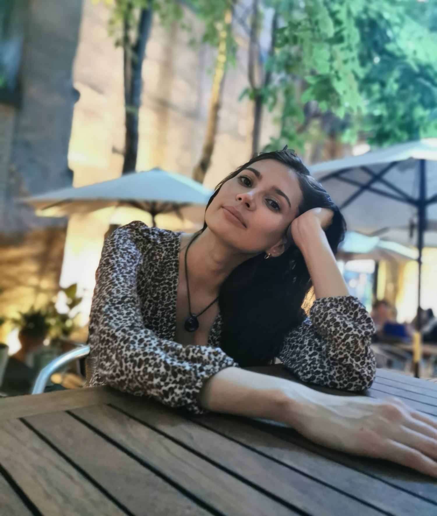 Woman in a leopard print top sitting at an outdoor table with greenery and buildings in the background.