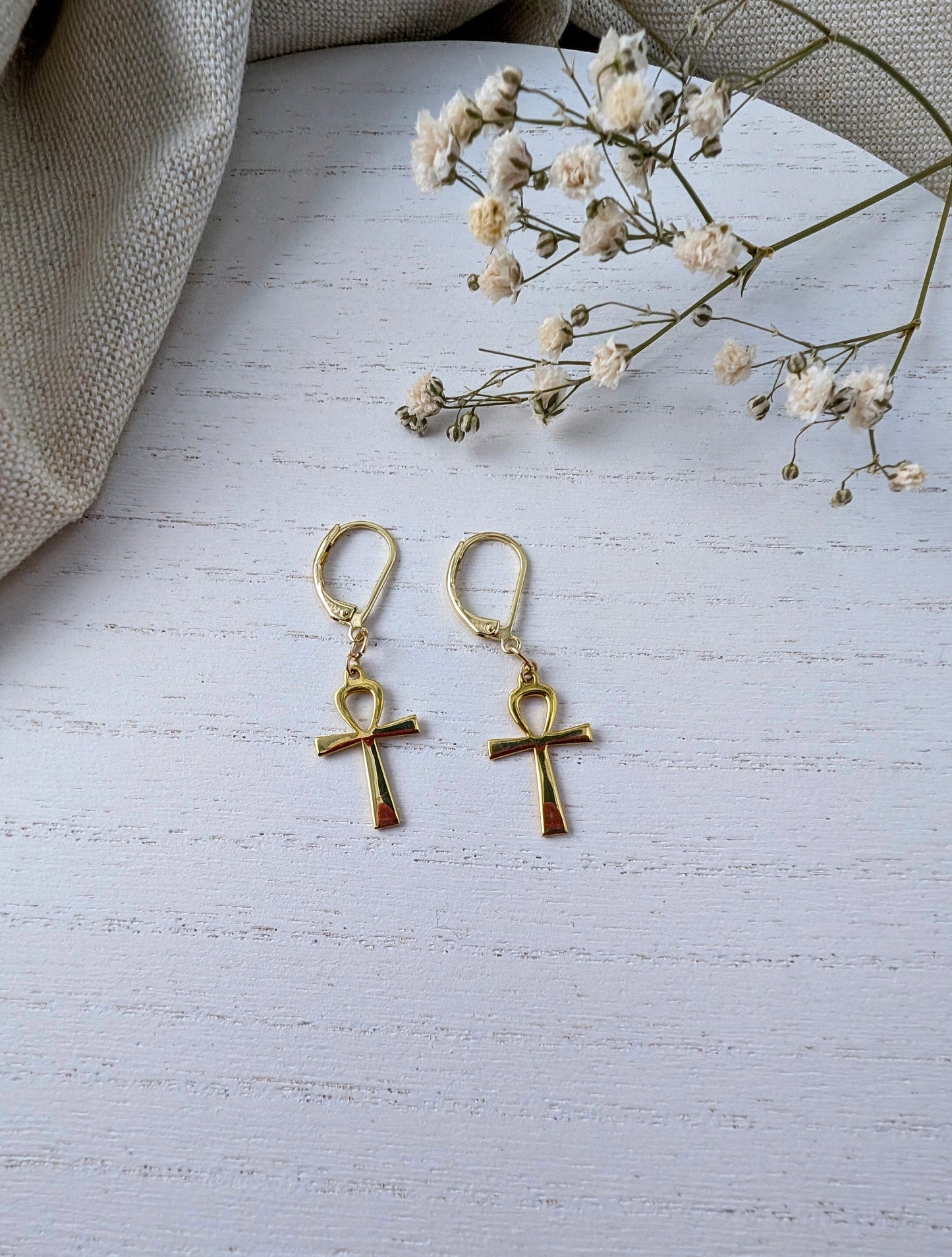 Gold ankh earrings on a light wooden surface with small white flowers.