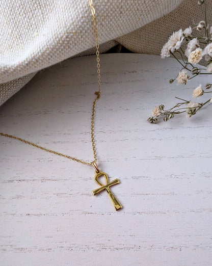 Gold necklace with an ankh pendant on a light wooden surface with small flowers.