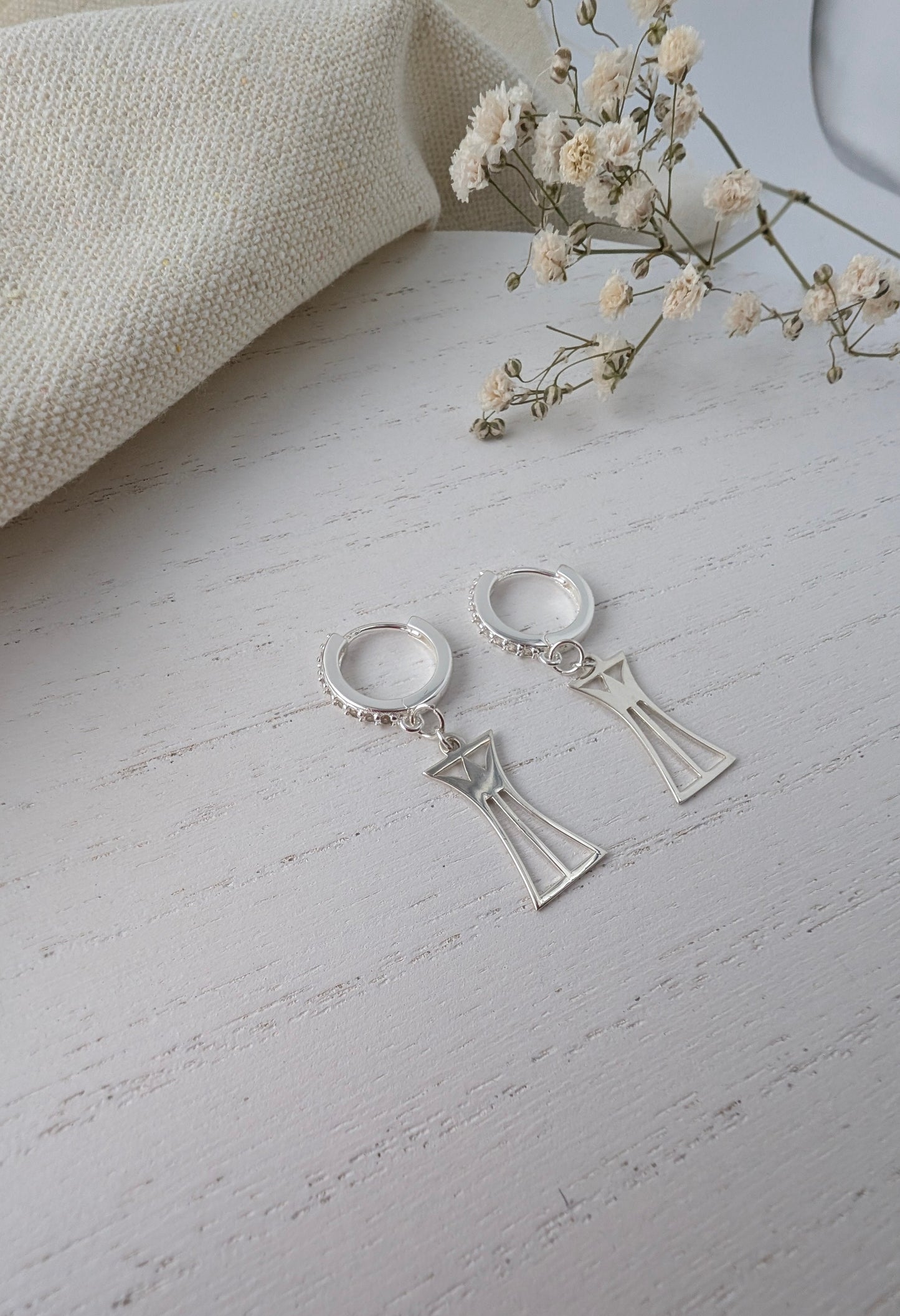 Silver lotus column earrings on a light gray surface with a beige fabric and small flowers in the background.