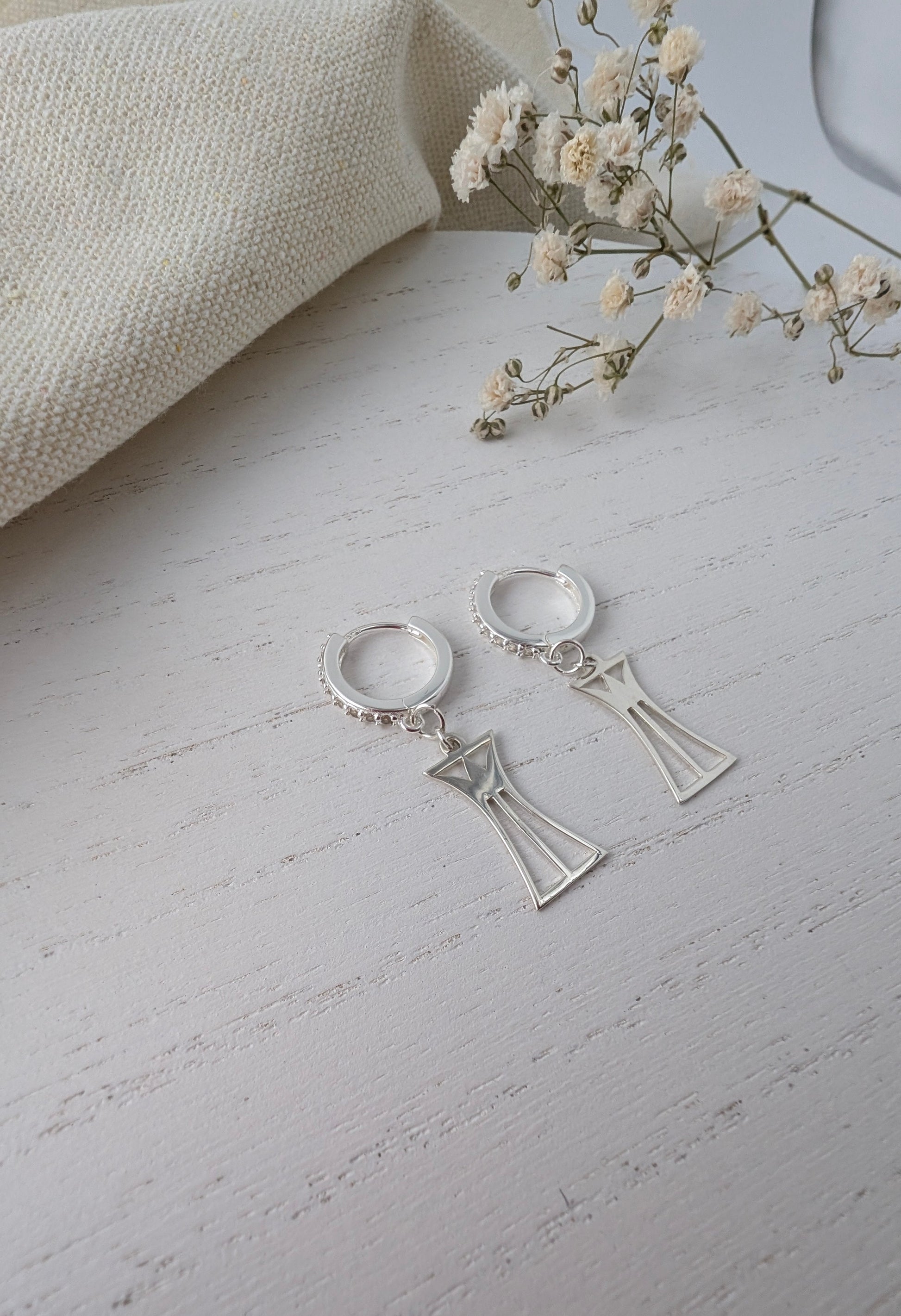 Silver lotus column earrings on a light gray surface with a beige fabric and small flowers in the background.