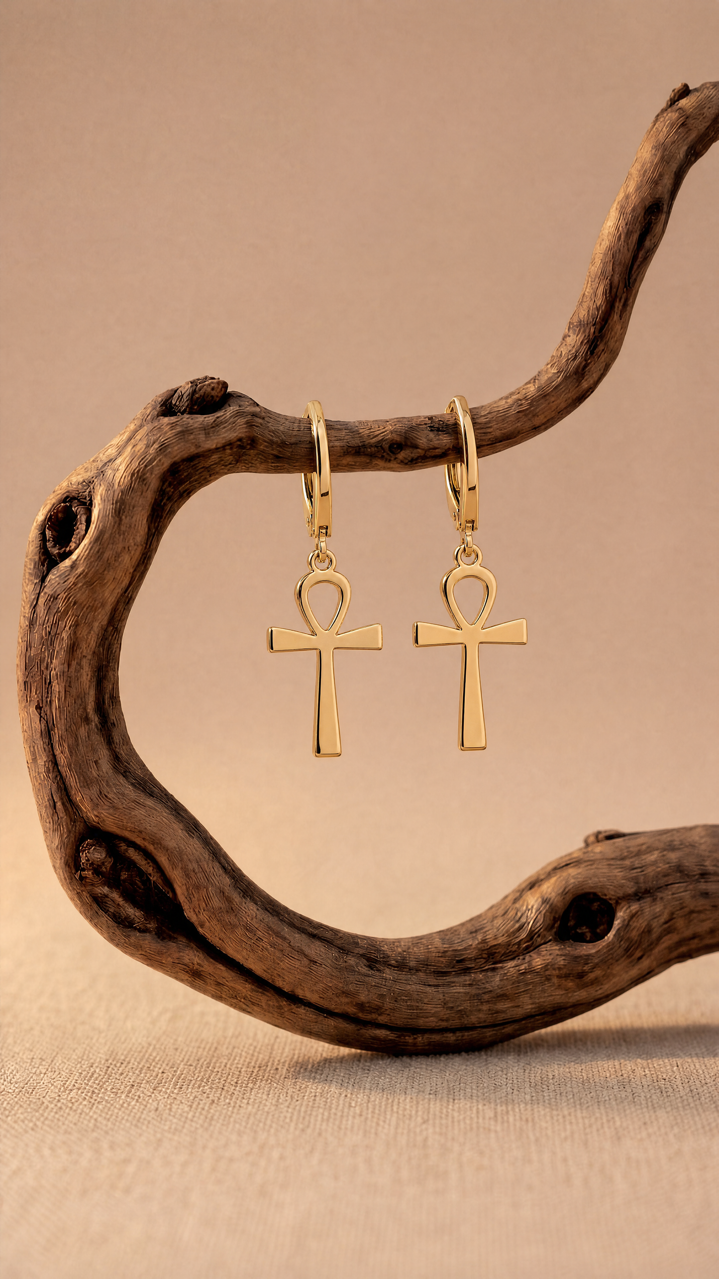 Gold ankh earrings on a wooden branch against a beige background