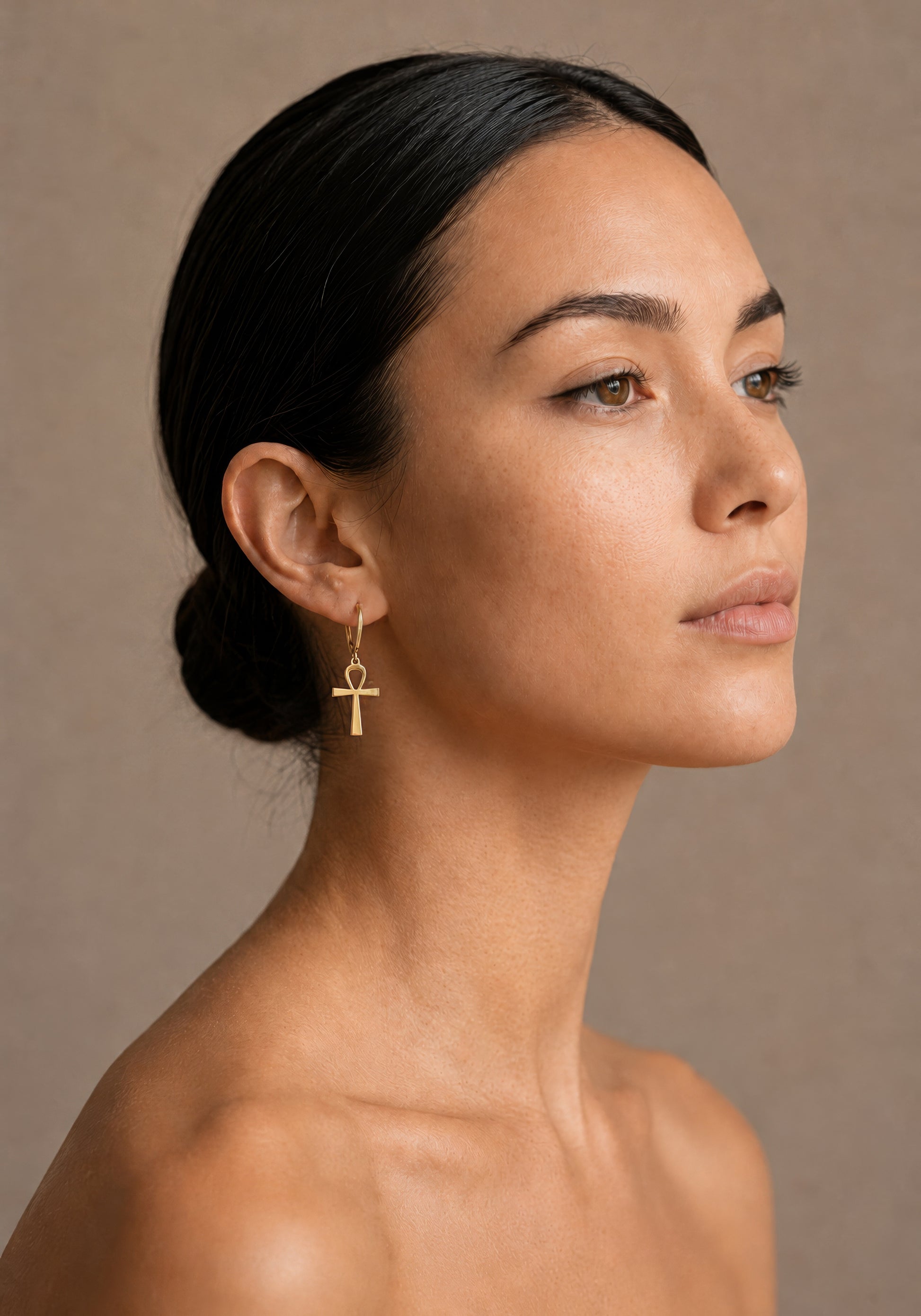 Woman wearing a gold earring with an ankh design against a neutral background