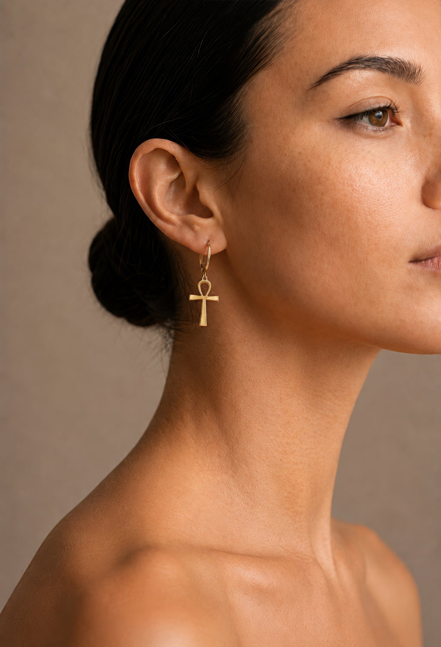 Close-up of a woman wearing a gold earring with ankh design against a neutral background