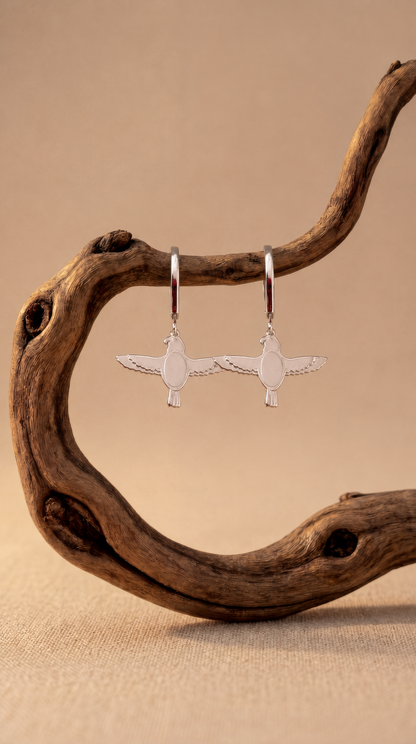 Silver falcon-shaped earrings hanging from a piece of driftwood on a beige background