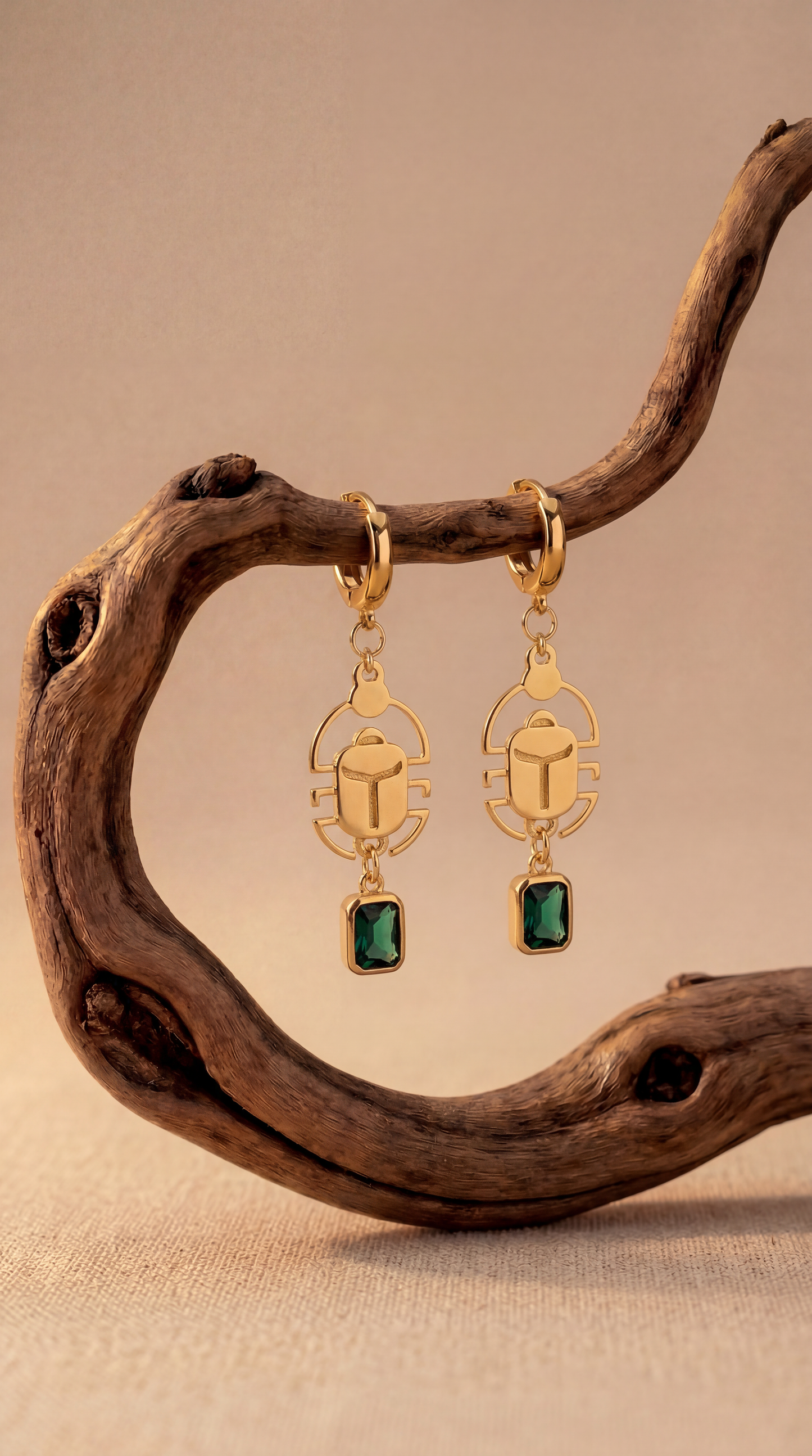 Gold scarab earrings with green gemstones on a wooden branch against a beige background