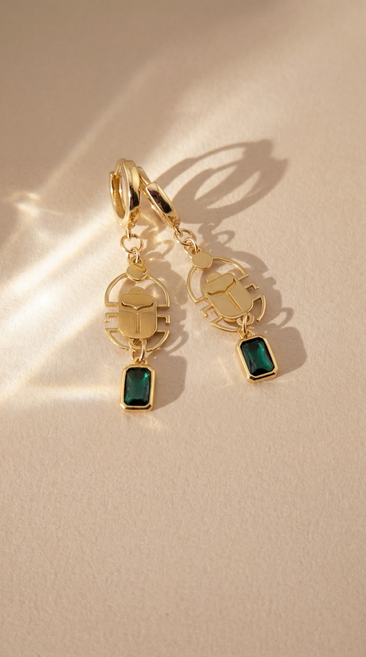 Gold scarab earrings with green gemstones on a beige background