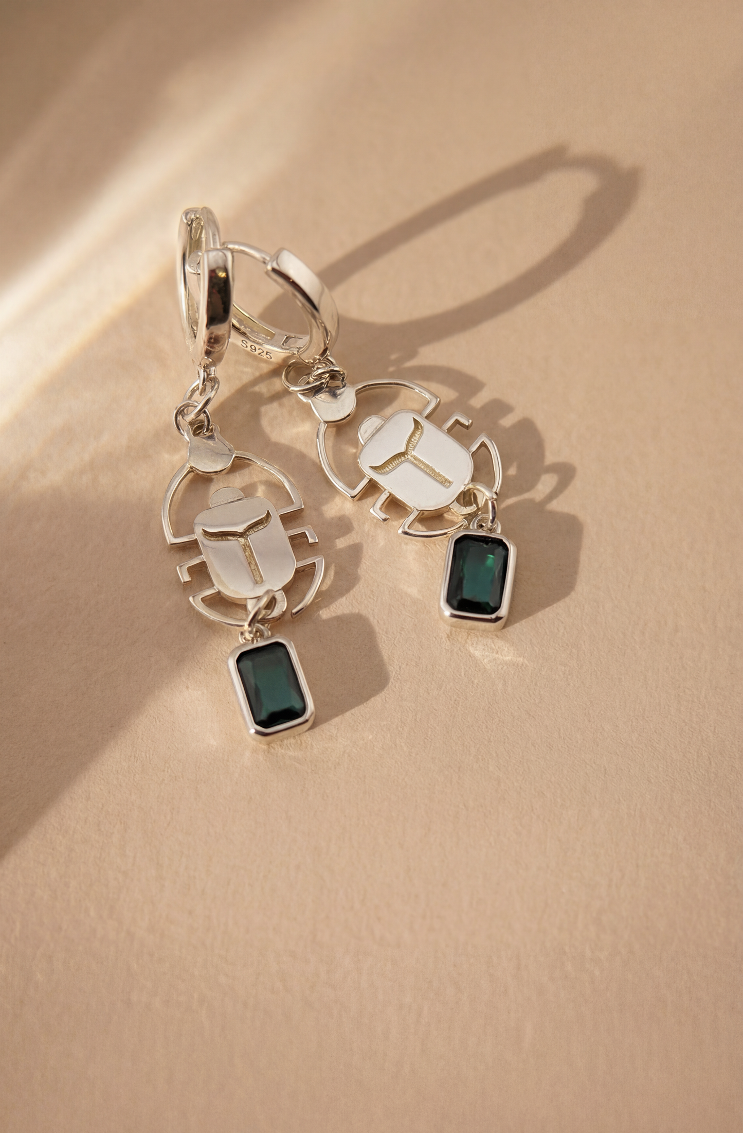Silver scarab earrings with green gemstones on a beige background