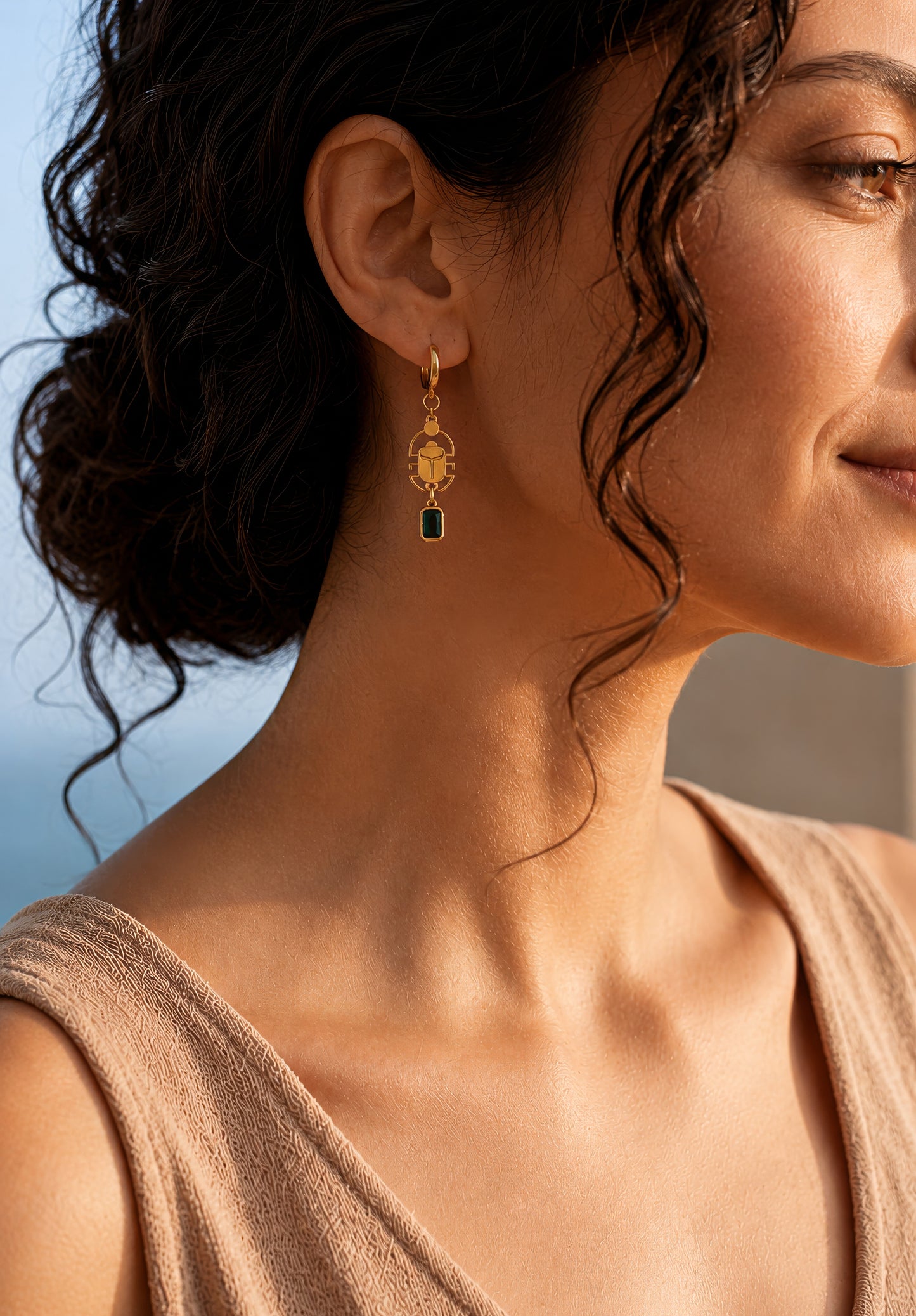 Close-up of a woman wearing gold scarabearrings with a green gemstone against a blurred outdoor background.
