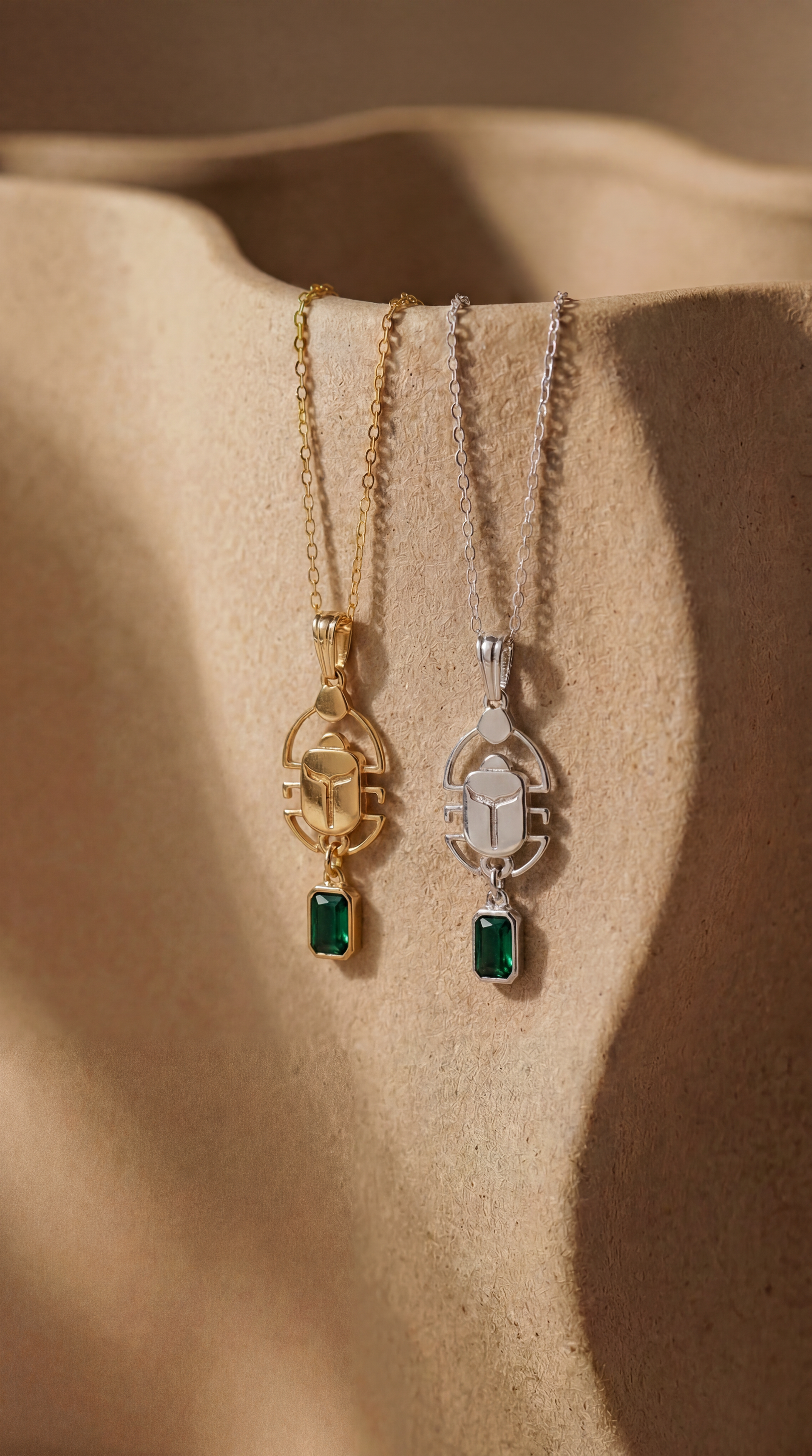 Gold and silver scarab necklaces with green gemstones on a beige ceramic surface